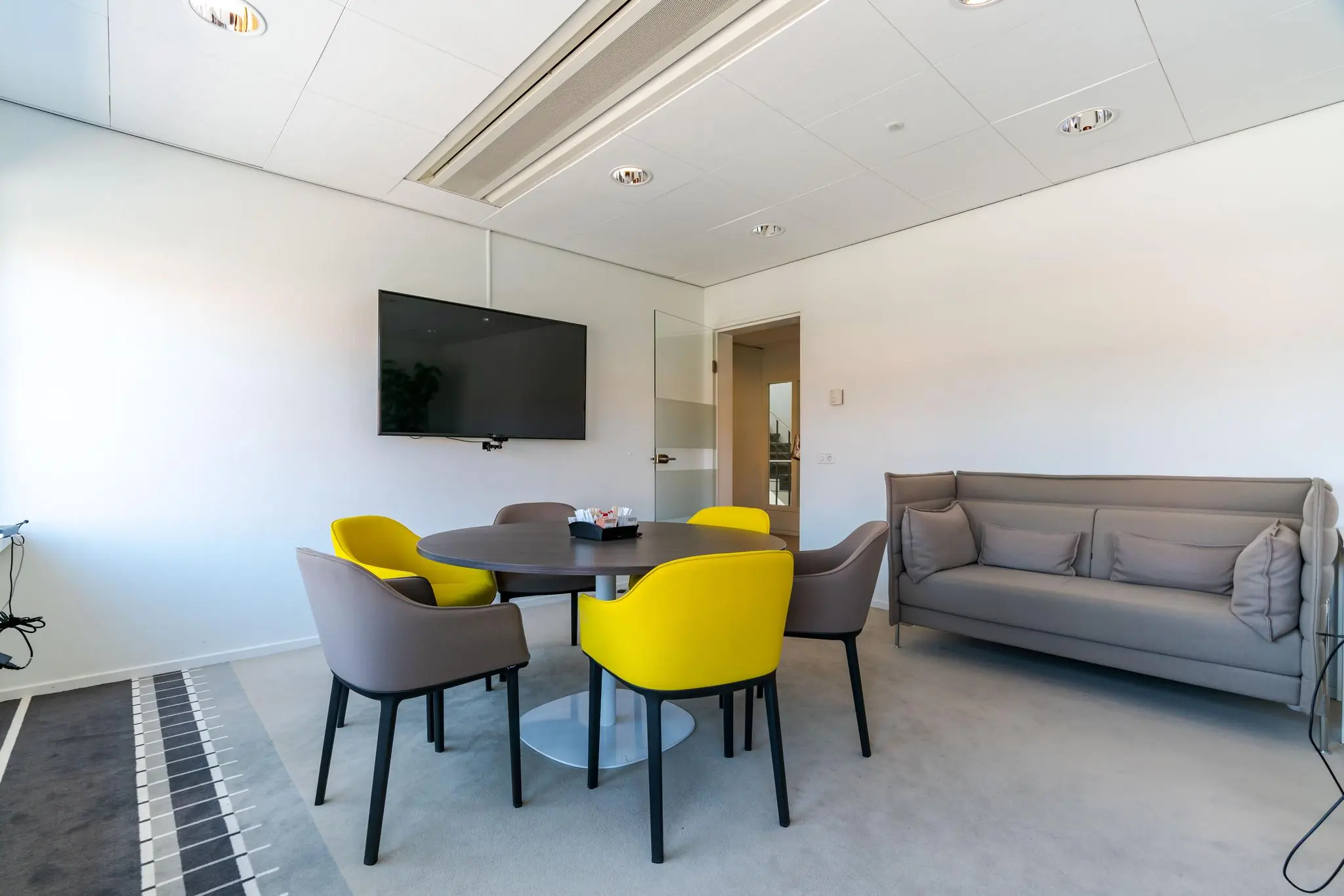 Moderne vergaderruimte aan de Van Alkemadelaan met ronde tafel, geel-grijze stoelen, grijze bank en flatscreen-tv aan de muur.